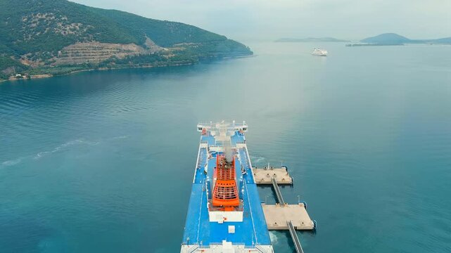 Igoumenitsa, Greece. Drone footage of a large blue-and-white ferry with an orange funnel departing from the port, with green coastal hills rising from the calm Ionian Sea under hazy summer light. Dro