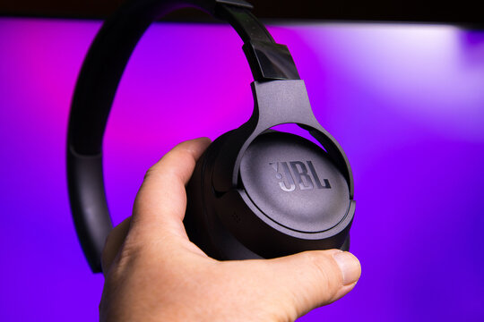 A close-up image featuring a hand holding JBL wireless over-ear headphones against a vibrant purple and black gradient background. The headphones showcase sleek black matte ear cups with the JBL logo