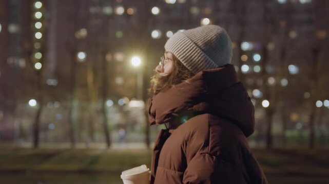 City night woman walking with coffee cup, passing pedestrians under glowing street lights, bundled in thick coat and beanie, occasional glance at skyline, subtle smile suggesting hopeful mood,