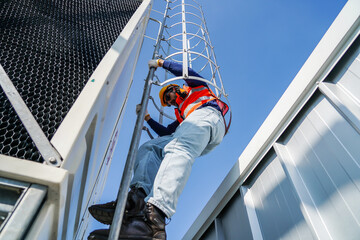 Male industrial technician in safety gear climbing a caged steel ladder on a commercial cooling tower against a clear blue sky. Maintenance, inspection, and occupational safety at high altitude. © TeTe Song