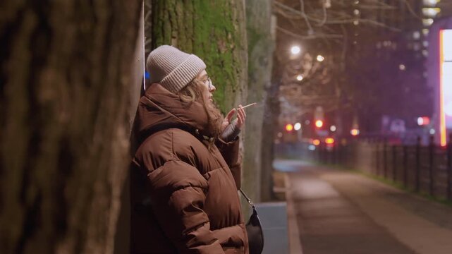 Young woman smoking beside tree. Nighttime street scene of solitary figure leaning against mossy trunk on sidewalk, wearing warm jacket and knit hat, cigarette in hand, slow exhale drifting into cold