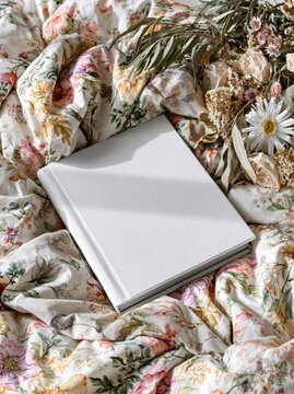 Blank white book resting on a floral patterned duvet with dried flowers and soft sunlight