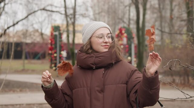 White woman with beanie examining leaves by tree trunk, calm botanist vibe, closeup on leaf texture, gentle breeze, soft smile, wearing brown puffer and glasses, study of seasonal decay, overcast