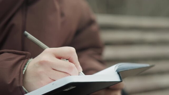 Closeup hands writing in notebook on bench, brown coat, silver pen, overcast light, reflective mood freelance writer drafting notes, steady strokes, page texture visible, shallow depth of field