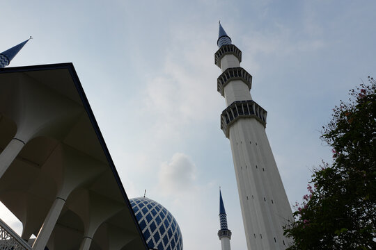 The grand minaret of the Sultan Salahuddin Abdul Aziz Mosque under a cloudy blue sky.