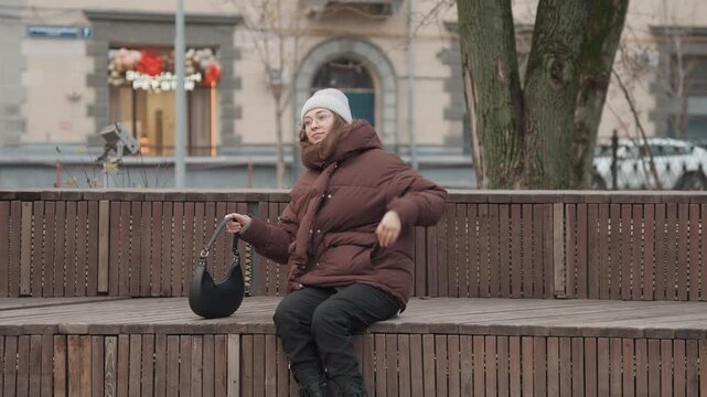 Caucasian woman scooter rider collapses on bench, wearing puffer jacket and beanie, clutching helmet before fainting empty plaza with wooden bench, leafless tree and muted winter light, scene conveys