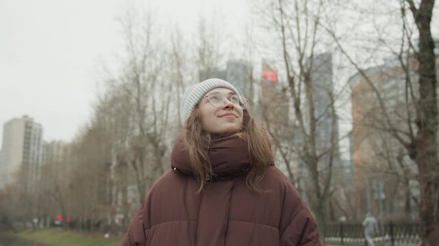 White woman looking up skyline, urban photographer pause in winter park wearing gray beanie and brown puffer coat with glasses, skyscrapers and bare trees in background, overcast sky and reflective