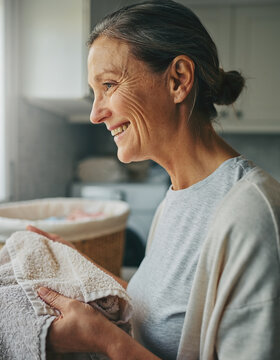 Smiling middle-aged woman folding laundry with towels inside bright kitchen