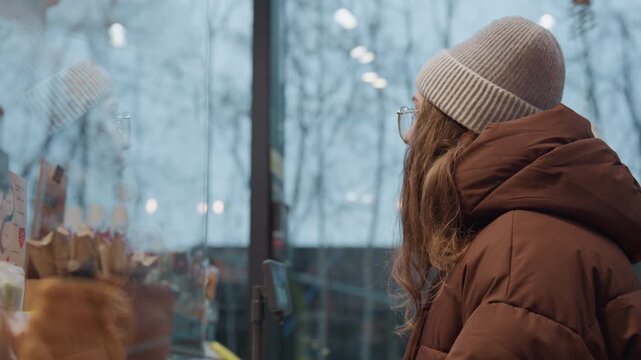 Contemplative white woman at winter window gazing through caf glass at pastry display and market lights, wearing knit beanie and puffer jacket, bare trees and soft bokeh outside frames suggest window