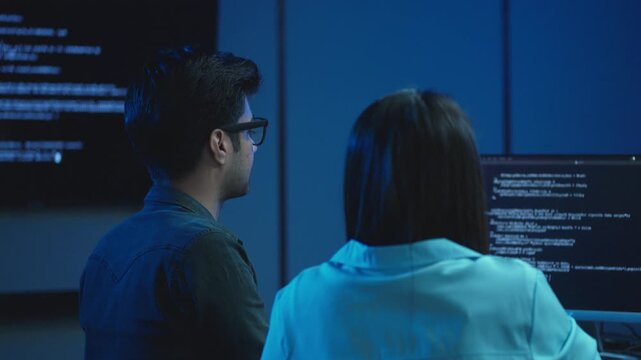 Two young software developers discuss program code displayed on a monitor while pointing at the screen in a dark blue lit modern office during a late night work session.