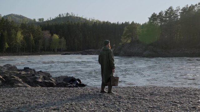 A man in a long protective raincoat stands with a bucket in his hand on the bank of a mountain river, then he falls poisoned by polluted water. An environmental disaster.