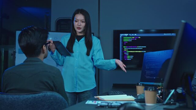 Young female developer points at code on a monitor while explaining a project to a male colleague in a dark blue lit modern technology office during night work.