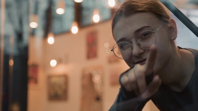 Intimate window portrait young woman smiling, wearing round glasses, leaning on cafe glass, warm string lights and soft bokeh in background, gentle finger point and heart gesture, cozy evening mood