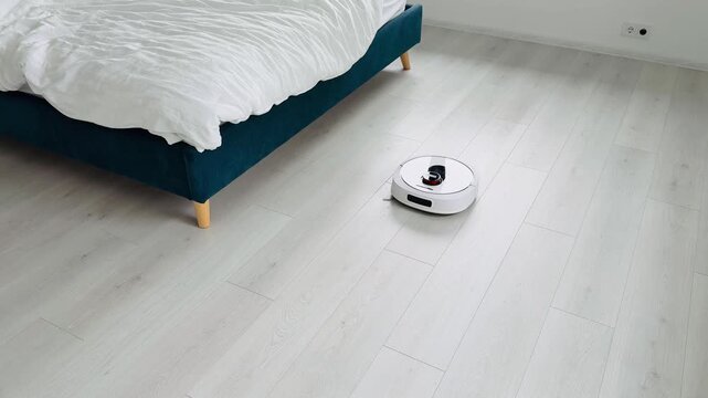 Robotic vacuum cleaner on light-colored wooden floor near modern bed with white bedding