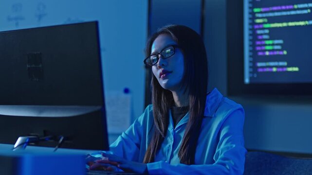 Focused young Asian female software engineer wearing glasses looks seriously at her computer monitor while debugging code and fixing programming errors in a tech office.