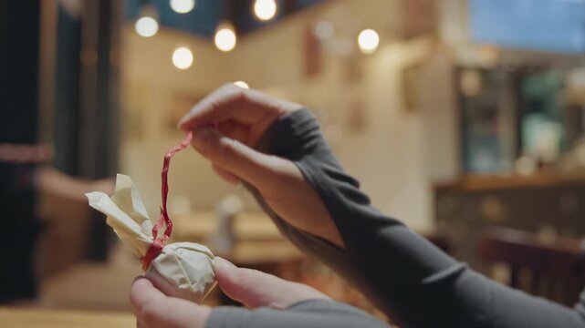 Hands opening tiny package over coffee cup in busy cafe, woman reveals treat with gentle smile, warm foam cup in foreground and cozy bokeh create intimate cinematic closeup of savoring small reward