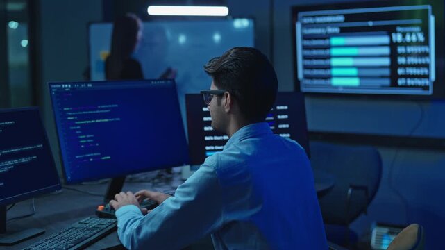 Young male software developer works at his computer surrounded by multiple code and data monitors in a dark blue lit modern technology office during night work.