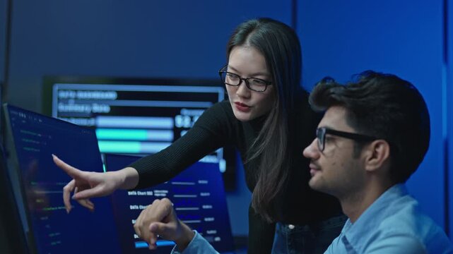 Young female developer points at code on a monitor while reviewing a project with a male colleague wearing glasses in a dark blue lit modern technology office.