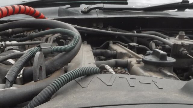 Closeup engine bay hoses wiring, mechanic inspecting engine during maintenance in garage, dusty surfaces and oilstained cover, red battery cable and corrugated tubing, metal intake manifold and clamps