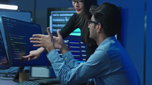 Male developer gestures while discussing a coding problem with a female colleague at a desk with code monitors in a dark blue lit modern technology office at night.