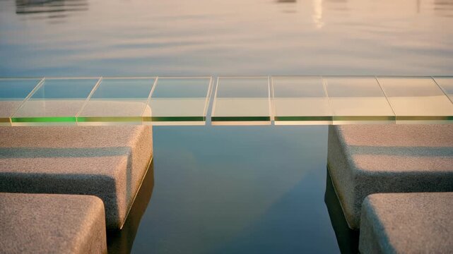 Transparent glass bridge over calm water between concrete pillars at sunset