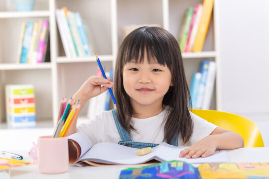 A polite and happy little Asian girl sitting up straight at her desk with crossed arms and smiling brightly at the camera.