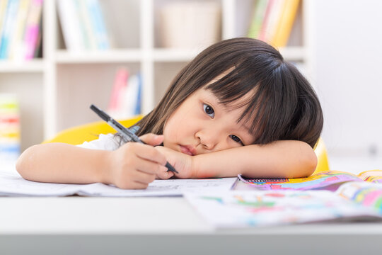 A tired little schoolgirl leaning her head on her arms on a desk while holding a pen during a boring study session.