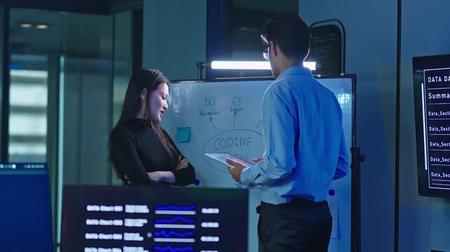 Male developer holding a tablet brainstorms with a female colleague at a whiteboard with coding diagrams in a dark blue lit modern technology office during night work.