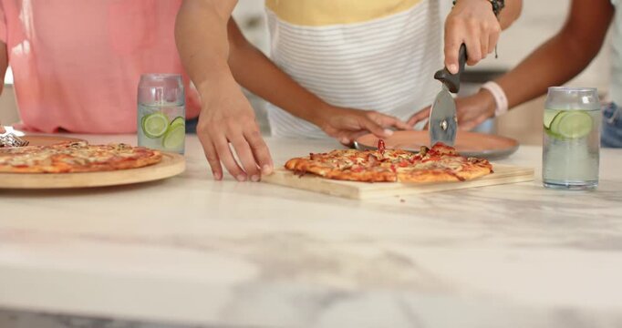 African American friends in kitchen hungry slicing pizza with pizza cutter, plating slices to share