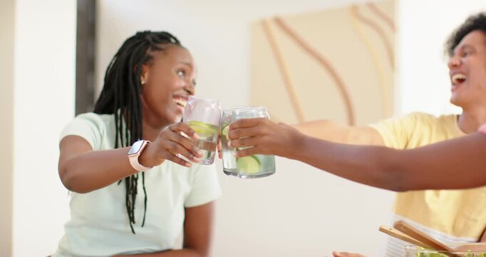 African American women clinking glasses with cucumber at table, urging third woman joining, sipping