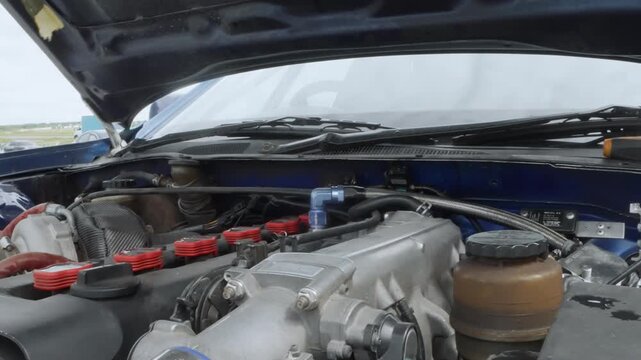 Engine bay inspection under pro hood, panoramic view of polished intake and braided hoses, prop rod holding hood, coolant reservoir and strut brace visible, pit crew performing methodical prerace