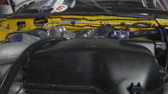 Closeup engine bay prerace inspection black intake cover and purple billet cap, yellow firewall accents, silicone hoses and intercooler piping, braided lines and compressor housing visible, daylight