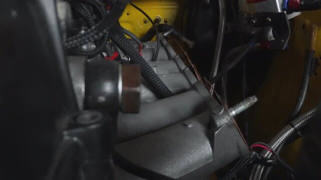Closeup engine bay braided wiring hoses, dark workshop lighting, black plastic airbox, silver intake tube, yellow painted engine block, exposed electrical connectors, threaded studs, textured rubber
