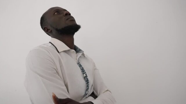 Hopeful man in white shirt looking. Black man gazes upward imagining romantic evening, patient suitor waiting for date subtle chest tilt, arms folded, minimal studio backdrop, quiet daydream mood