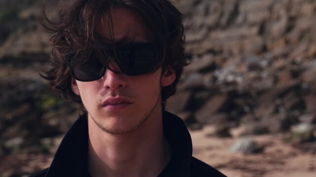 Calm Young man with brown hair blown on wind against rock on summer sunny day. Attractive young man looks at the camera on the nature. Handsome guy in black sunglasses poses outdoors. Slow motion