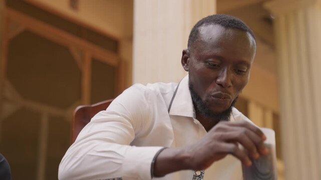Black man preparing for romantic date, seated indoors sipping water and collecting thoughts, white shirt, calm expression and polite smile, quiet courtyard atmosphere, gentle lighting.