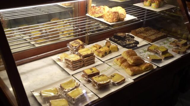 Assorted bakery pastries, cakes, croissants, and packaged bread displayed in a warm glass showcase for cafe and bakery shop content.