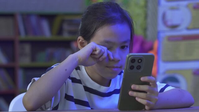 Little Asian girl rubbing her eyes while using a smartphone, suffering from digital eye strain.
