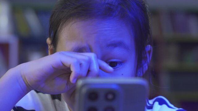 Little Asian girl rubbing her eyes while using a smartphone, suffering from digital eye strain.