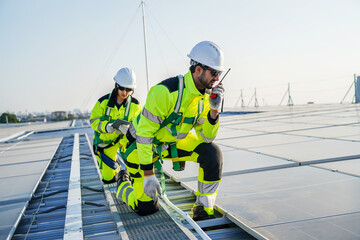 Two professional engineers in safety gear inspecting solar panels on a roof. A male worker uses a radio while a female technician checks data on a tablet. Renewable green energy industry concept. © TeTe Song