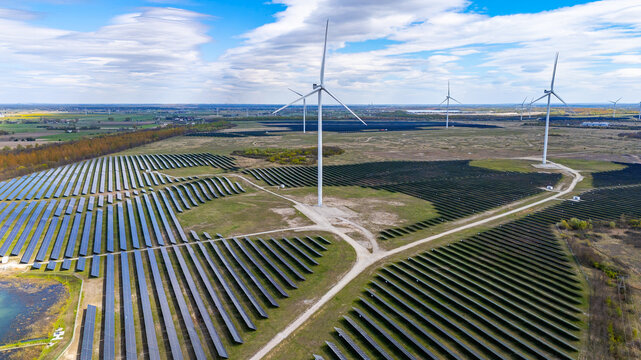 Sunny, panoramic aerial drone view of a hybrid renewable energy farm featuring rows of blue solar panels and modern white wind turbines against green fields, a blue sky, and scattered clouds.
