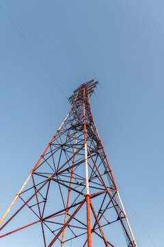 Tall telecommunications tower with red and white structure reaching into a clear blue sky, showcasing metal framework and power lines extending from the top
