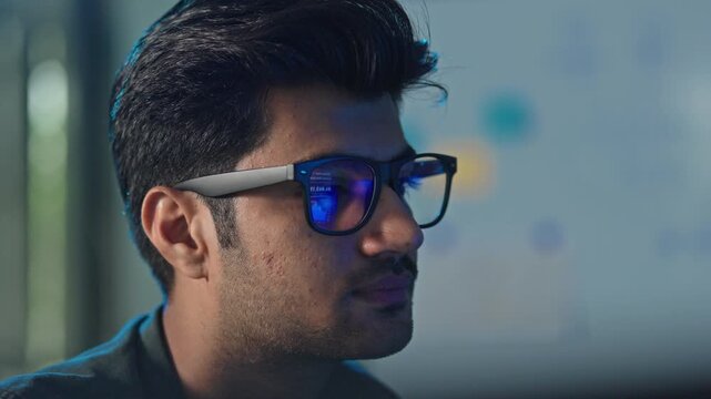 Close up profile of a young male software developer wearing glasses with screen light reflecting on his face in a bright modern technology office workspace at work.