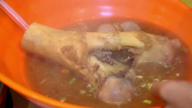 Steaming beef bone soup in an orange bowl, featuring rich broth, tender meat, and meatballs in a close-up street food serving scene.