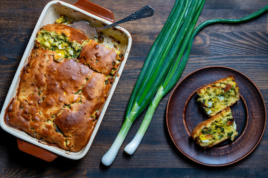 Homemade scallion and egg pie on dark wooden table, closeup. Top view of a savory baked pie with green onions and boiled eggs in a baking dish and slices on a clay plate