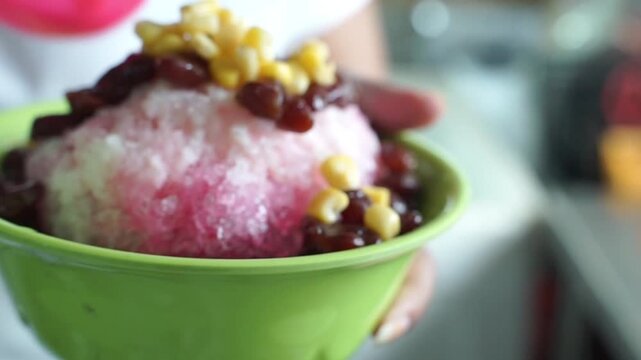 Colorful shaved ice dessert topped with sweet corn, red beans, and jelly pearls in a green bowl, a refreshing Asian street food treat.