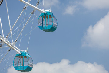 Two fully enclosed gondola cabins hang from a Ferris wheel's white support beams, set against soft fluffy clouds on a clear blue sky, embodying concepts of leisure as well as aspiration and freedom. © thomasgweber