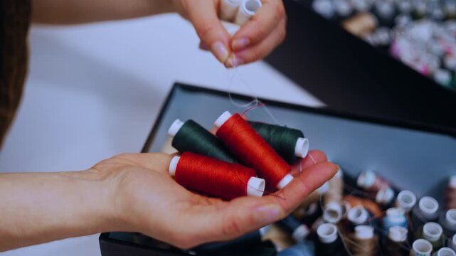 Female hands browse a thread assortment box filled with dozens of spools, then pick a red bobbin. The camera focuses on her fingertips as they sort through the rainbow of options with purpose and care