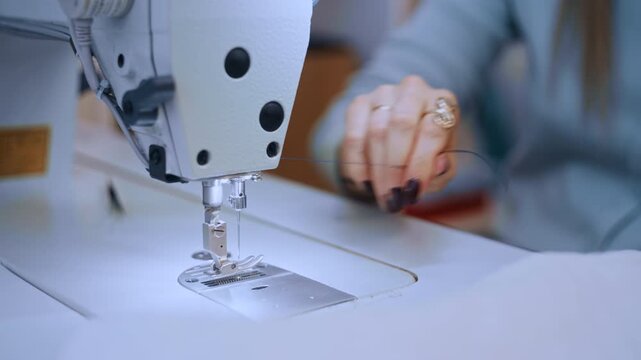 A close-up shot of a woman's hands threading the needle of a sewing machine in a tailor's workshop. She squints with concentration, holding the thread taut before pushing it through the small opening