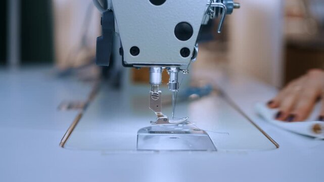 An unidentified factory worker operates an industrial overlock machine on white textile. Her fingers keep the fabric aligned while the machine's blades cut away excess material